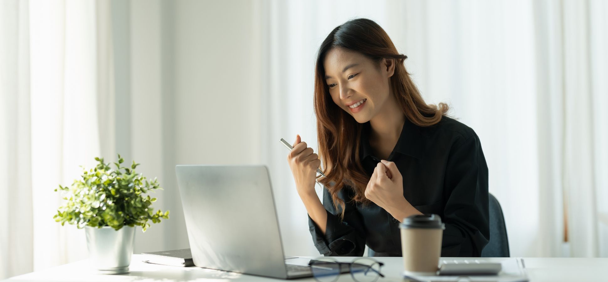 A young woman in a black shirt smiles confidently while working on a laptop in a bright, modern office. She holds a pen in one hand and raises both fists in a gesture of success or excitement. A coffee cup, notebook, glasses, and a small potted plant sit on the white desk in front of her.