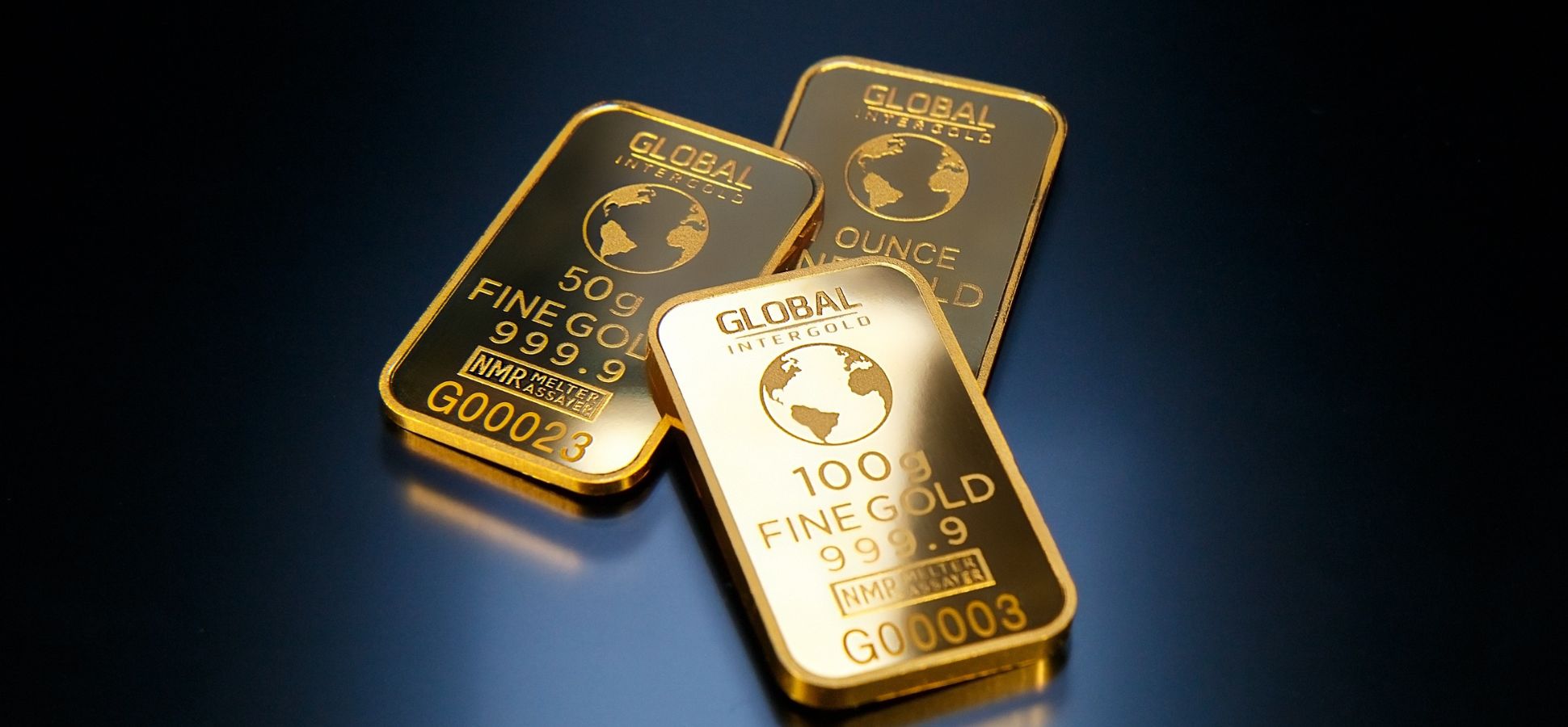 Three Global Intergold fine gold bars, weighing 50g, 100g, and one ounce, are arranged on a dark, reflective surface.