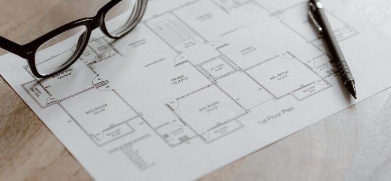Black-framed eyeglasses and a pen resting on a printed architectural floor plan on a wooden desk