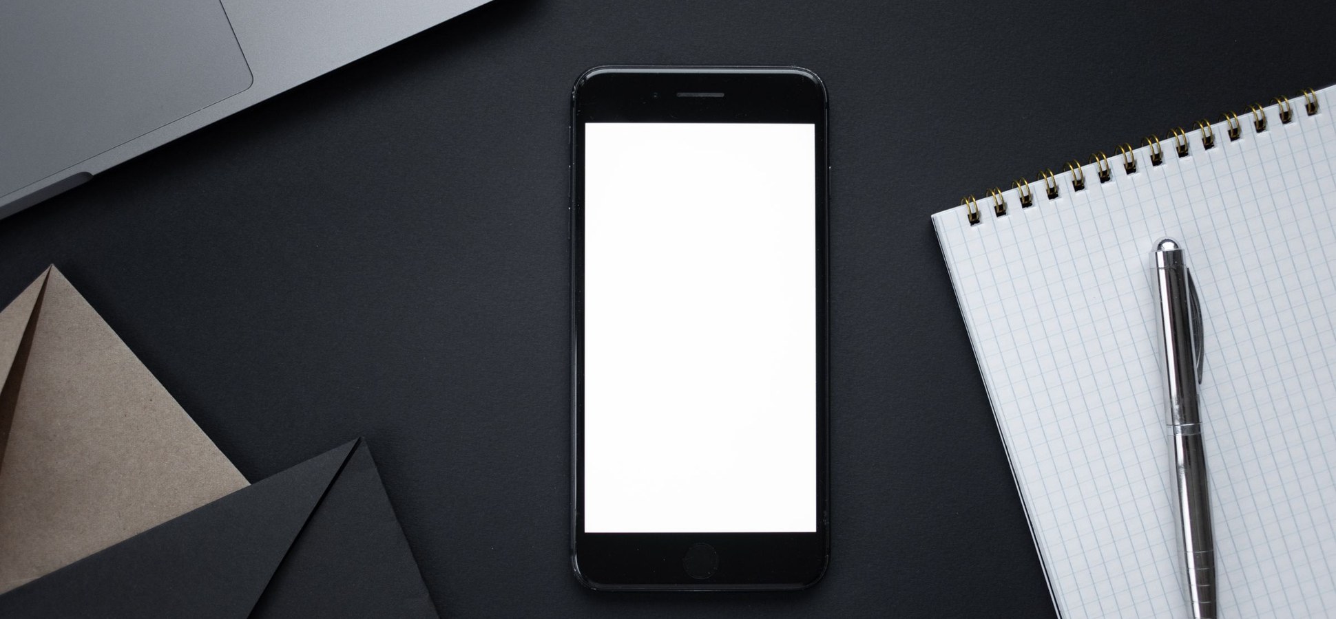 A smartphone with a blank screen beside a notebook and pen on a dark desk.