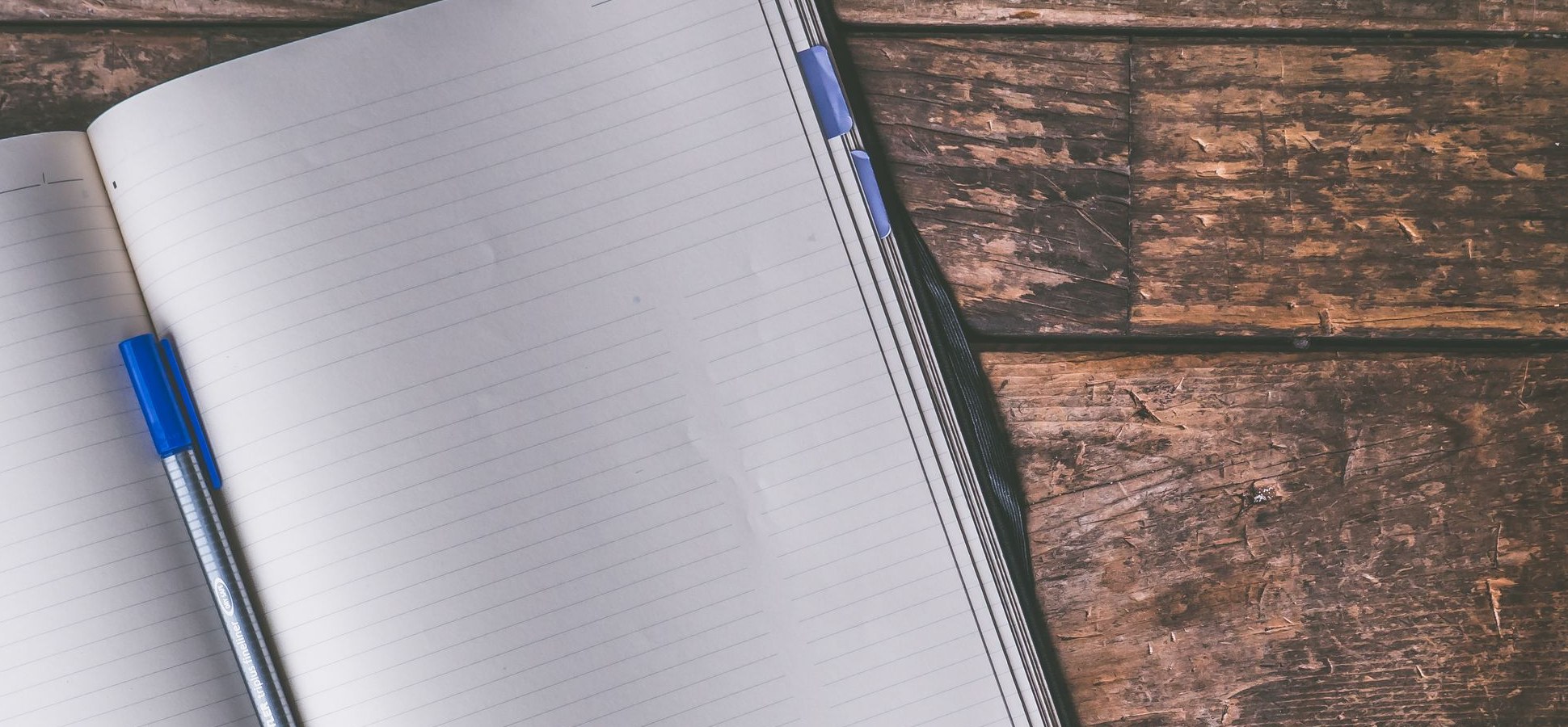 An open blank lined notebook with a blue pen on a wooden table.